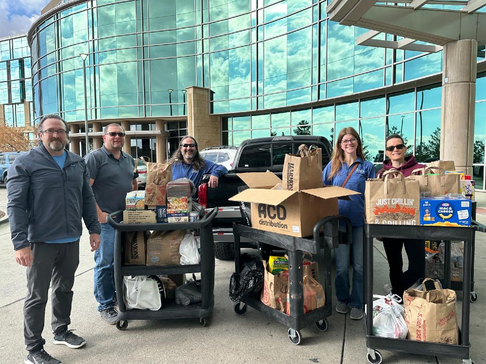 Five people stand outside a building with large glass windows, smiling and posing with carts full of assorted groceries and supplies.