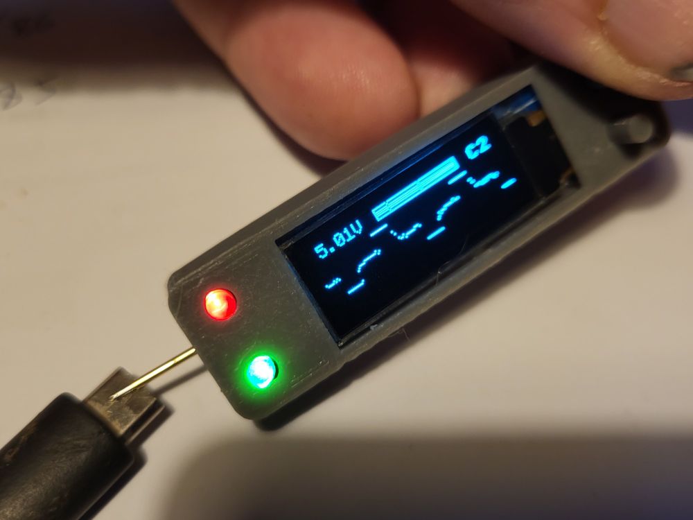 Small grey 3d printed box held between thumb and forefinger against a white desktop showing 5.1v and a squared off AC waveform. Red and green leds are lit at one end. 