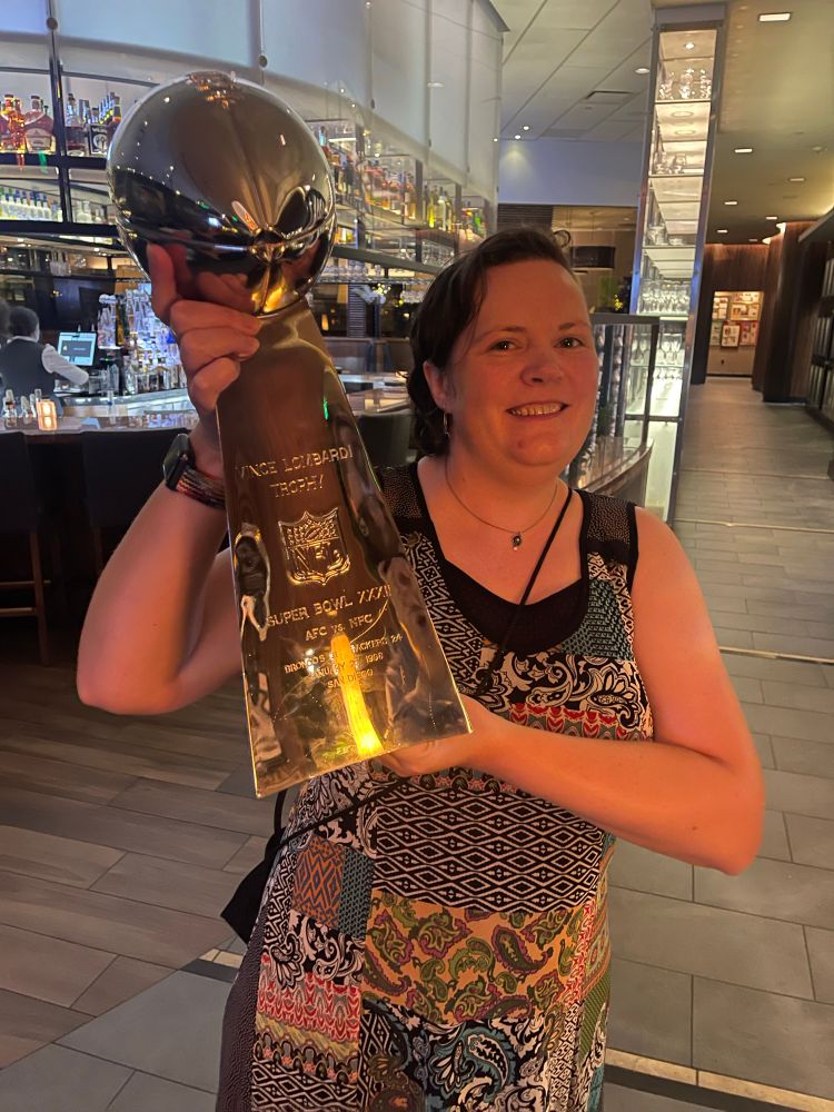 A pale woman in a dress grins like an idiot while holding a large Lombardi trophy. 