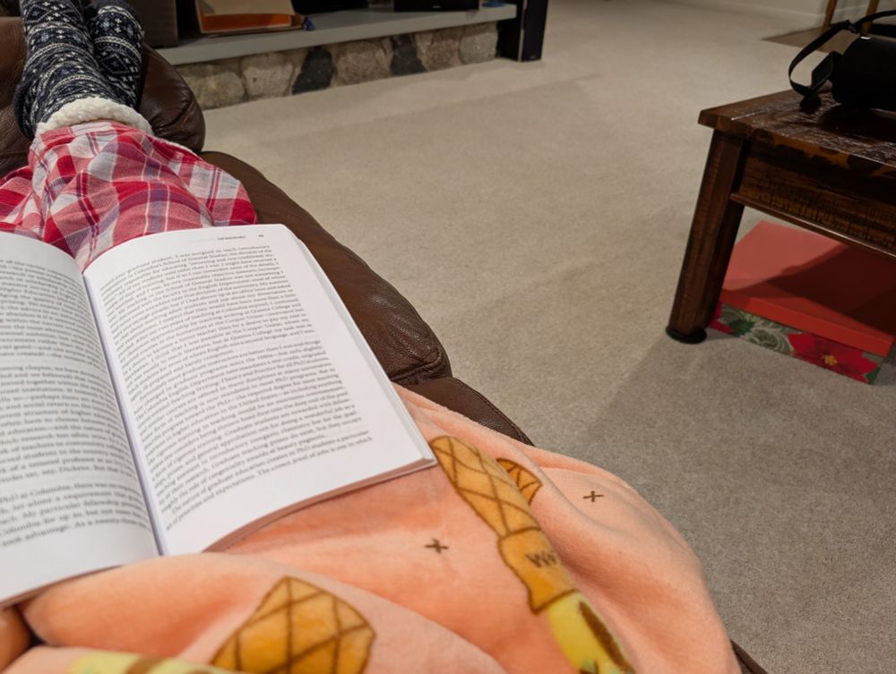 Picture of me in my living room, reading a book. I am wearing cozy slipper socks, red plaid pajama pants, and a big oversized fluffy hoodie with drawings of ice cream cones on it.