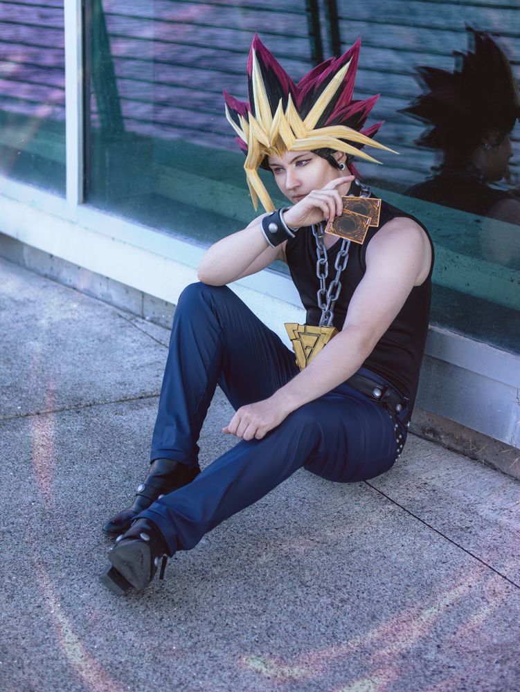 Yami Yugi siting against glass, he holds cards up obscuring part of his face. 