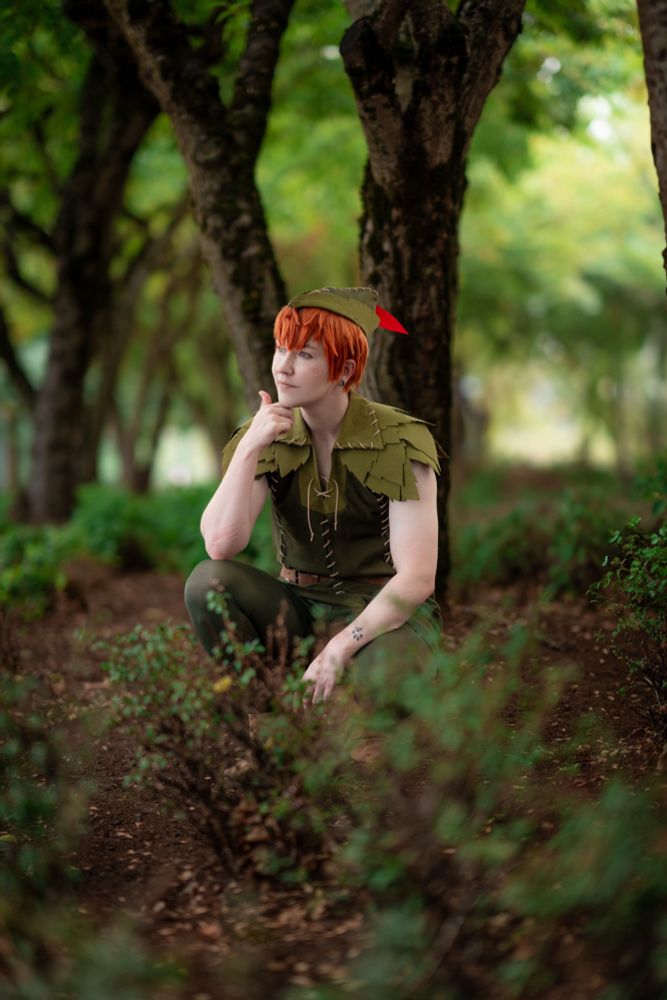 Cosplayer dressed as Peter Pan crouches in a forest area with bushes all around. There are a number of trees in the background. He has one arm raised to his chin thoughtfully. He wears a green tennis with fabric leaves across his shoulders. He has a matching hat with a red feather. 