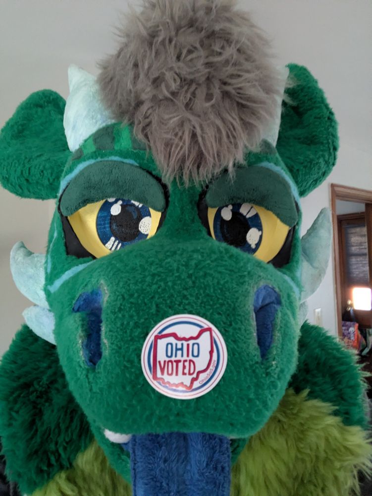 A green dragon fursuit head with an Ohio "I Voted" sticker on the snout.
