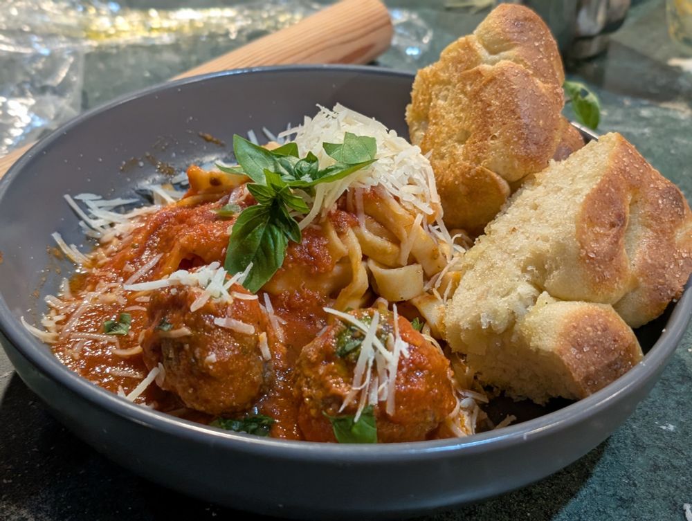a plate of wide pasta noodles and meatballs with fluffy bread, furnished with herbs and cheese