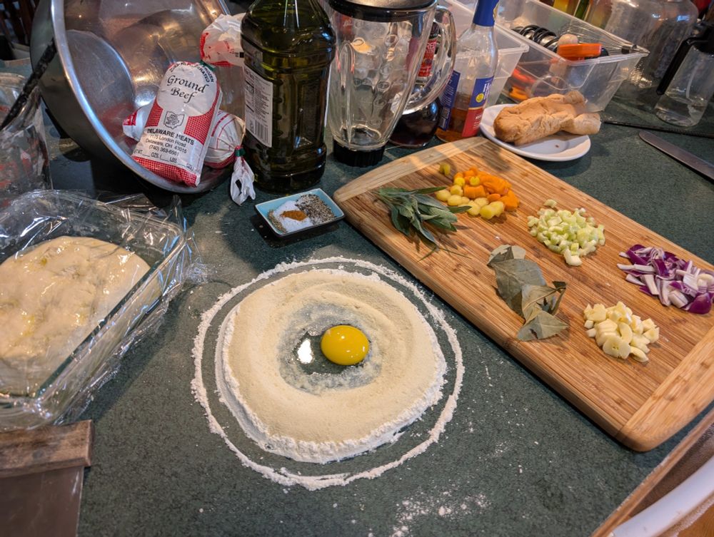 a collection of ingredients set out and ready to make a meal including a circle of two kinds of flour with an egg and some water in the middle of it, like a noodle pyramid. there is also some bread dough in a pan to the left chopped up vegetables including carrot celery and onion and garlic laid out on the cutting board and some spices and 2 lb of ground beef and a bunch of other stuff