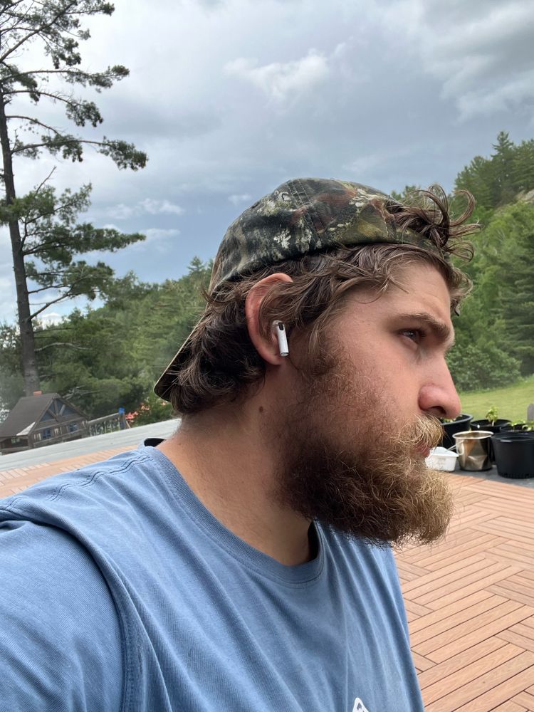 Bushy bearded man looking to the right with , AirPods in, bushy rugged hair peeking through and around a backwards camo hat. His beard is extending past his nose and moustache connects into beard, lightest shade of brown, with his underchin beard and behind his ears being the darkest shade of brown. Overcast day in the background with planters, large white pine in the left corner and red pines and other trees going up a hill to the right. Appears to be standing on a deck with a smaller structure in the back left, looking forward with piercing dark eyes and a furrowed brow, some frown lines appearing above the brow, wearing a light blue shirt but can’t see past his breasts
