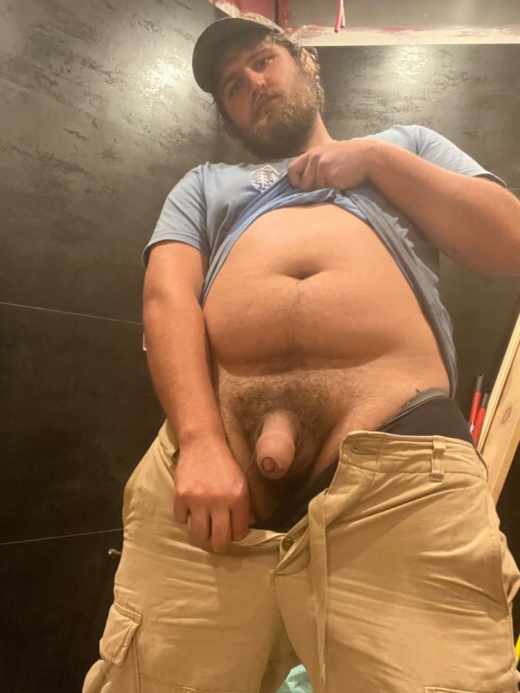 Hairy bearded man on his lunch break in the washroom with a mop in the background he’s standing centre frame with legs apart. One hand is pulling his tan pants and black underwear down to reveal a semi hard/ wide as it is tall uncut cock dangling above a ballsac of the same size and nestled in a thick hairy bush of pubes. His other hand is holding up his shirt halfway exposing his belly and it’s casting a shadow where it overhangs above the genitals where the bush is following up to the belly button with a “happy trail”. He’s got an expectant expression with a slight frown “gesturing”.
The caption says “who’s on cleanup duty?”