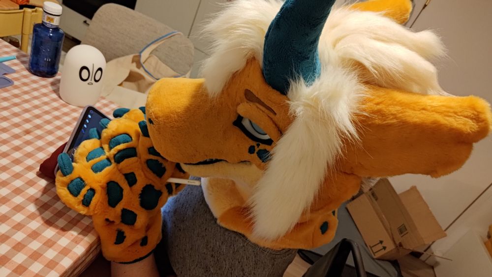 Foto de mi en picado desde el lado, llevando mi cabeza y manos de fursuit de mi fursona Enyard mientras estoy dibujando en mi iPad. 

En esta la cara de mi cabeza de fursuit tiene los párpados de manera que parezco cansado. En la mesa tengo al lado una lámpara de Napstablook de Undertale apagada. 