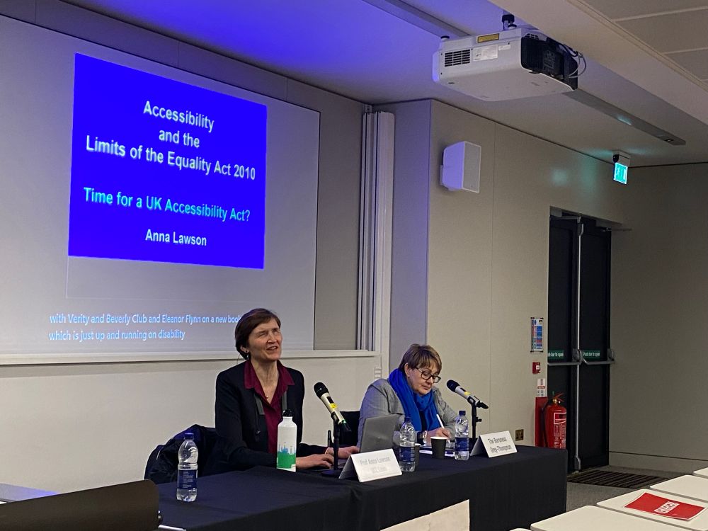 Anna Lawson sits next to Baroness Grey-Thompson with microphones in front of them. Behind them a blue PowerPoint slide that reads “Accessibility and the Limits of the Equality Act 2010. Time for a UK Accessibility Act?”