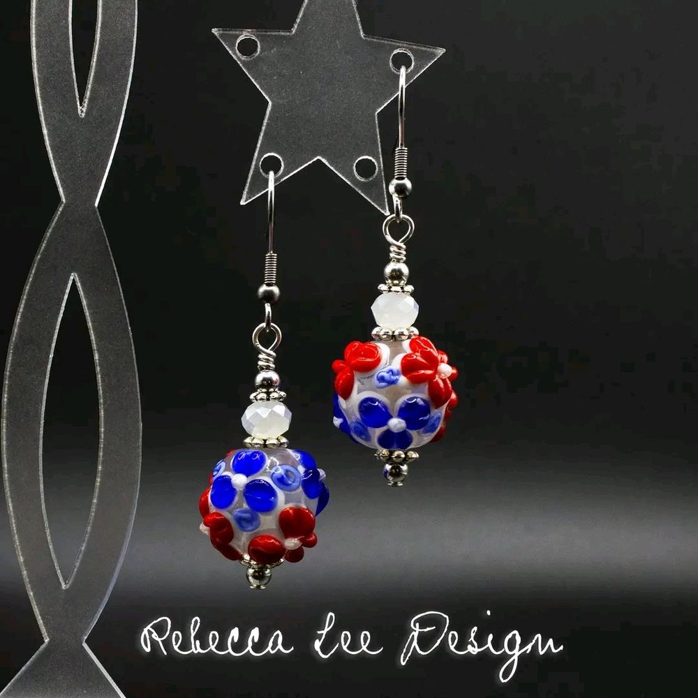 A pair of dangling earrings made with red, white and blue floral lampwork glass beads, white faceted crystals, and silver colored metals. The earrings are hanging from a clear stand against a black background. The bottom of the picture is signed "Rebecca Lee Design."