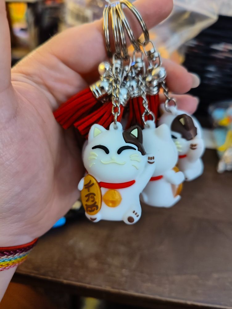 Three cute beckoning lucky cat (maneki neko) keychains hanging in my palm. The front cat is facing the camera while the others are facing away. Each cat is mostly white but has a brown spot on their left ear. The inside ear triangles are pale yellow. They have happy eyes-closed expressions with black eye lines and pale yellow nose, mouth, and whiskers. They're wearing red collars with large gold bells. They have brown paw prints on their feet and their raised left "hand". They hold a gold oval plaque with Japanese writing in their right hand.

They're hanging from the key rings on short chains with red faux suede tassels and tiny bells at the top. The rings, chains, tassel caps, and bells are silver colored.