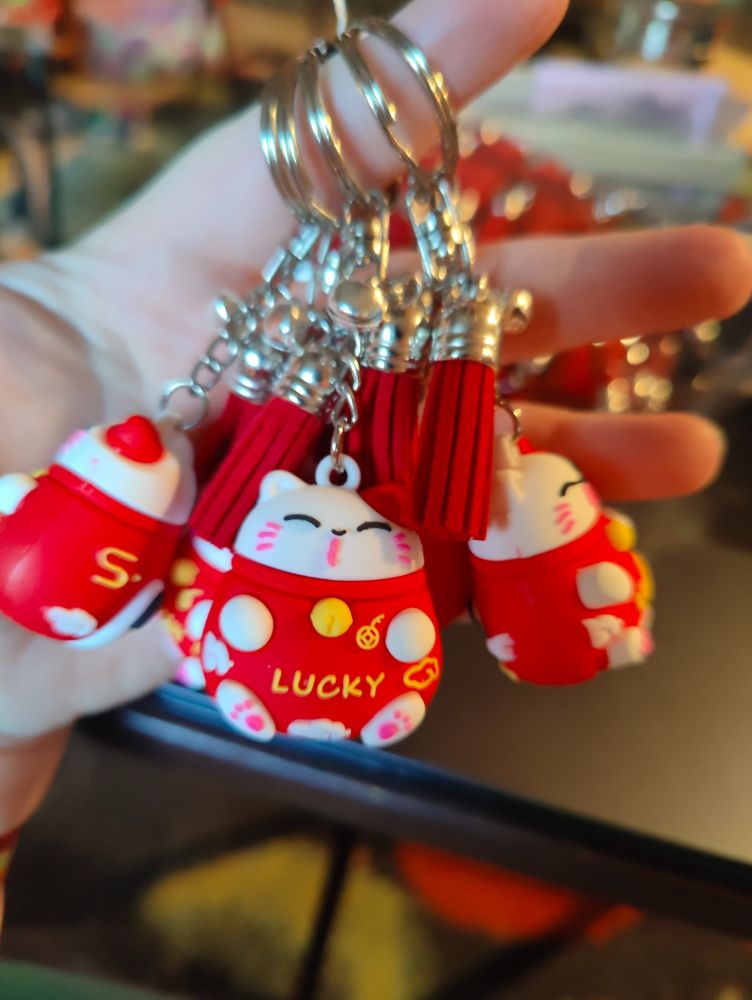 A handful of cute red and white lucky beckoning cat (maneki neko)  keychains. The one in front faces forward while the others face in different directions. 

The cats have white faces with a red left ear and white paws. They're wearing red collars with yellow bells, and red outfits on their rounded bodies. The word "Lucky" is written in yellow across their bellies and there are other small white and yellow designs on the sides and bottom of the body. They have pink paw prints on their feet and a pink "kissy" mouth, pink whiskers, and happy closed eyes lined with black and little black noses. 

They're hanging from the key rings on short chains with red faux suede tassels and tiny bells at the top. The rings, chains, tassel caps, and bells are silver colored.
