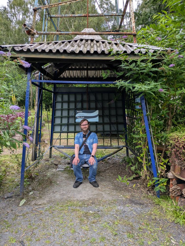 A photo of a guy in a blue shirt and jeans sat under a wind turbine with plants either side