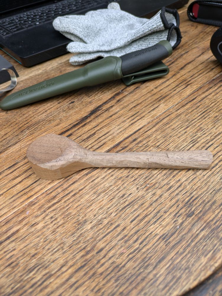 A photo of a wooden spoon under construction on an old wooden table with a sheathed knife and gloves in the background 