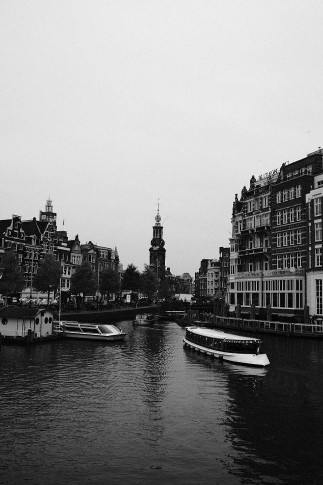 A canal in Amsterdam in the lower half of the image. Two boats are on it. The upper half has buildings and the sky. In the center is a tower pretty contrasty in front of the light sky. Left and right are houses.