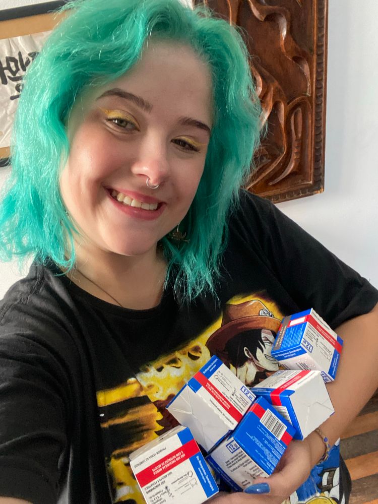 Eu, mulher branca com cabelo verde e olhos claros, com delineado amarelo e camiseta preta com os irmãos Ace, Sabo e Luffy de One Piece, segurando 5 caixas do medicamento Somatropina 