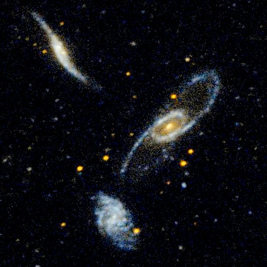 The frame is dominated by three spiral galaxies in shades of yellow and blue. The largest is at the center of the frame. The disk of the galaxy is tilted, so that it appears oval-shaped, with the long axis running diagonally from upper right to lower left. It has a bright yellow center, surrounded by a ring structure. Emerging from the ring are two spiral arms, defined by blue star clusters. The arms for a tiled, backward S-shape. At the bottom left is a smaller face-on spiral galaxy. Unlike the other two galaxies in the group, this galaxy does not appear distorted. It has a small core, surrounded by loose spiral arms that are filled with blue star clusters. At upper left is a distorted spiral galaxy seen nearly edge-on. Its disk is warped, forming a thin, nearly vertical, backward S-shape.