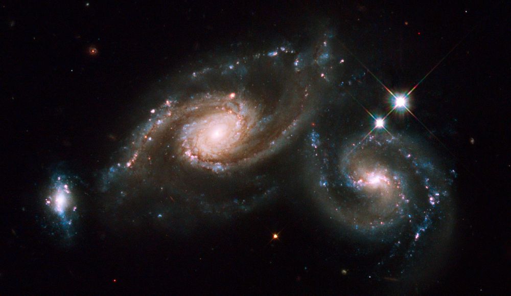 Three overlapping galaxies fill the frame. In the center are two large spiral galaxies. The larger one, at center-left, has a bright nucleus surrounded by a yellow-white core made of mostly older stars. At the edge of the core, two spiral arms emerge. One begins at about 6 o'clock and curves counter-clockwise to about 12 o'clock. The second begins at about 12 o'clock and curves down to about 8 o'clock, with a fainter part of the arm continuing on to about 5 o'clock. The arms are threaded with brown dust lanes. A few pink star-forming regions and blue star clusters dot the outer portion of the arm. The second spiral galaxy is at center-right. It has a bright center, and a well-defined bar and two spiral arms. Together, the bar and arms form an S-shape. Compared to its companion, this galaxy has less dust and more blue star clusters in its arms. The final galaxy in the triplet appears at the far left of the frame. It is much smaller than its companions and has a blobby, irregular appearance, although it could be a disrupted disk seen edge-on. It has a bright yellow-white center and an almost vertical line of blue star clusters. Dotted throughout the galaxy are pink star-forming regions. A few stars appear on the black background of space, including two large foreground stars, which appear above the center-right galaxy.