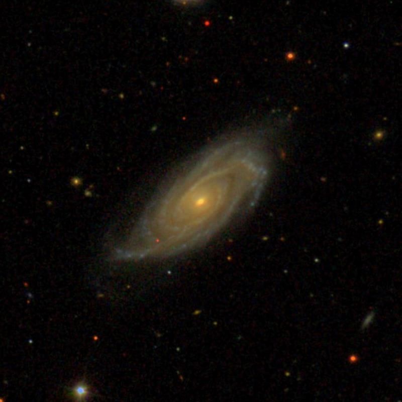 A spiral galaxy with a small, bright, circular core. The muted, yellowish-green spiral arms wind clockwise around the yellow core. The galaxy is tilted slightly, so it forms an oval shape, rather than a circle. In the background are scattered red, yellow and green dots, which are stars.
