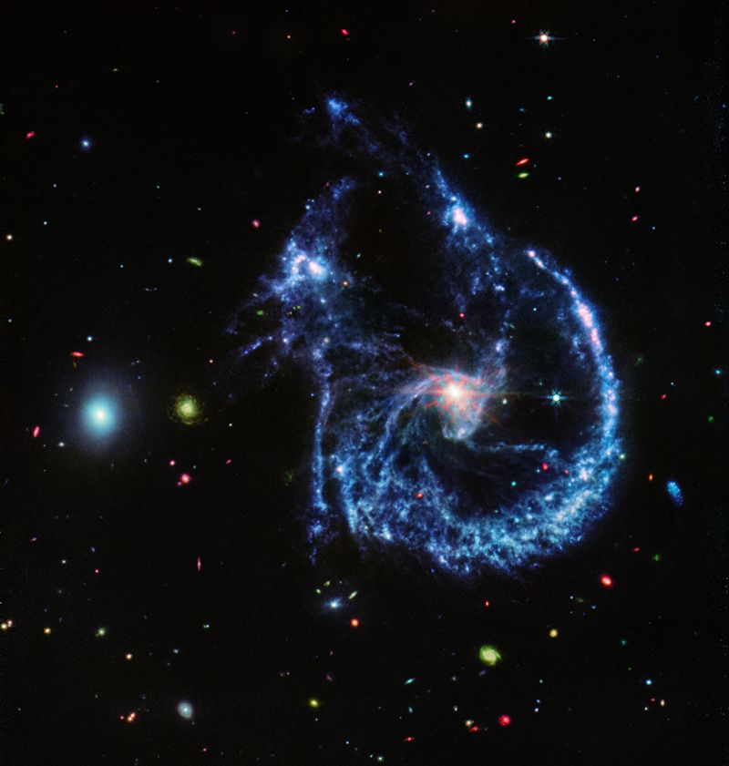 A pair of interacting galaxies. The larger of the two is slightly right of center. It is a one-armed spiral galaxy with a hazy, bright, white center and a ring of blue, gaseous filaments. The center of this galaxy shows JWST’s eight-pronged diffraction pattern. The single spiral arm begins at about the 7 o'clock position and spirals counterclockwise to the 12 o'clock position, forming a ring structure. Toward the bottom left and right of the ring are filaments of gas spiraling inward toward the core. At the top left of the ring is a noticeable gap, bordered by two large, blue pockets of dust and gas. The smaller galaxy is made of hazy, light blue gas and dust. Many red, green, blue, and yellow galaxies are spread throughout, with some being hazier in composition and others having more defined spiral patterns.