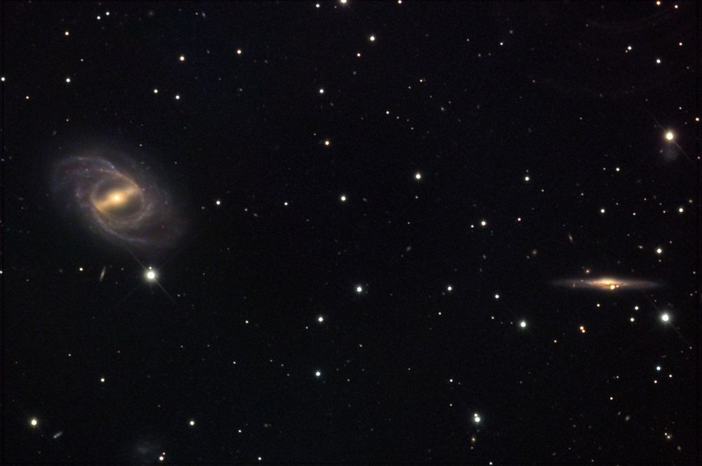 Two small galaxies appear on a sparse field of stars. At center left is NGC 2523, a face-on barred spiral. It has a bright yellow central core with a horizontal bar-shaped structure that connects to a thin ring. The area between the ring, core, and bar is dark. This central part looks like a steering wheel. The spiral arms originate at the ring and spiral outward counterclockwise. The arms are dotted by blue star clusters and shrouded in a haze of stars. At right center is NGC 2523B, an edge-on spiral.  It has a yellow central bulge and a blue disk, which is crossed by a central brown dust lane.