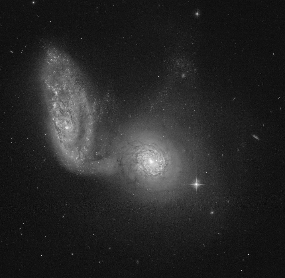 A pair of interacting spiral galaxies. The galaxy in the center appears face-on with a bright center, surrounded by swirls of dark dust filaments. The outer areas of the galaxy are filled with a haze of pale stars. To the left is its companion galaxy. The disk of this galaxy is seen at an angle, tilted so that it appears to be an oval rather than a circle. Dark dust filaments somewhat obscure the spiral structures in its disk, which is filled with star clusters. At the bottom left edge of this galaxy, the end of a spiral arm or a tidal tail appears to extend downwards, connecting to the side of its companion. At the top edge of the central galaxy is what appears to be a faint tidal tail, dotted with star clusters, perhaps a continuation of the tail from the companion galaxy. The black background of space is dotted with foreground stars and tiny background galaxies.