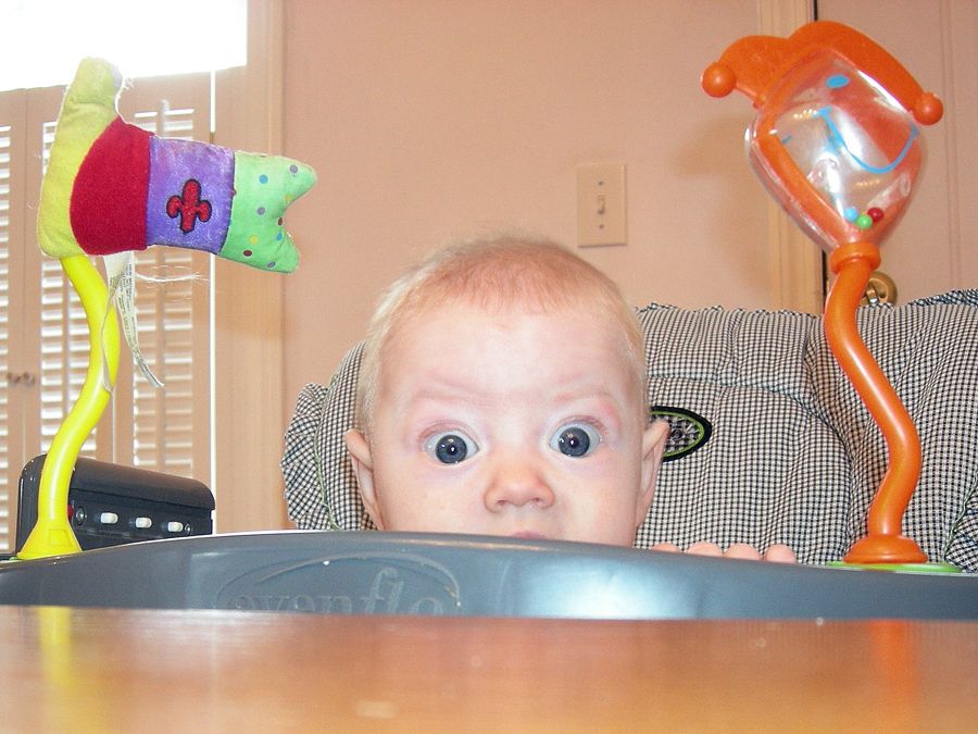 A baby is peeking above the tray of her high chair, framed by whimsical colorful toys on either side.  Her eyes are opened very wide and she looks hilariously surprised.