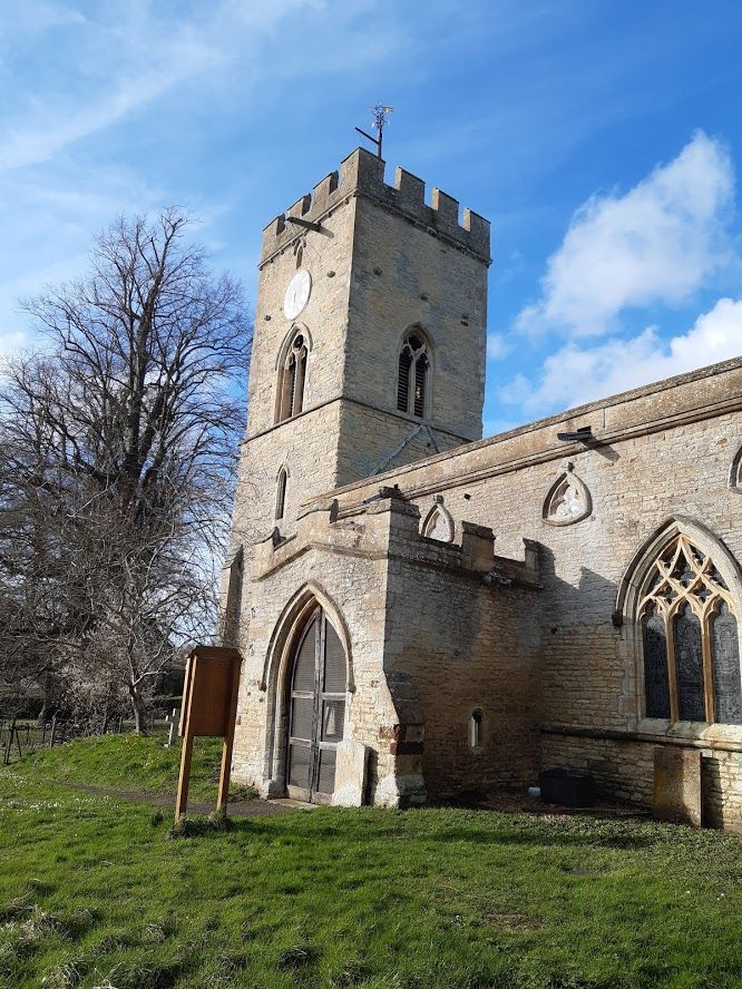 Against a blue sky with pale white clouds, the pale stone tower of Cranford St Andrew stands out. There are no leaves on the trees to the left and the chuchyard grass is green with hints of snowdrops. 
There's a crenelated parapet, and some very prominent rain spouts. There's a round clock that's glinting in the sun above louvres. On the side of the tower facing the nave there's evidence of a past higher roof line. The church has a large porch and attractive rounded triangular windows along in a line above the more conventional window we can see at the right of the image. 