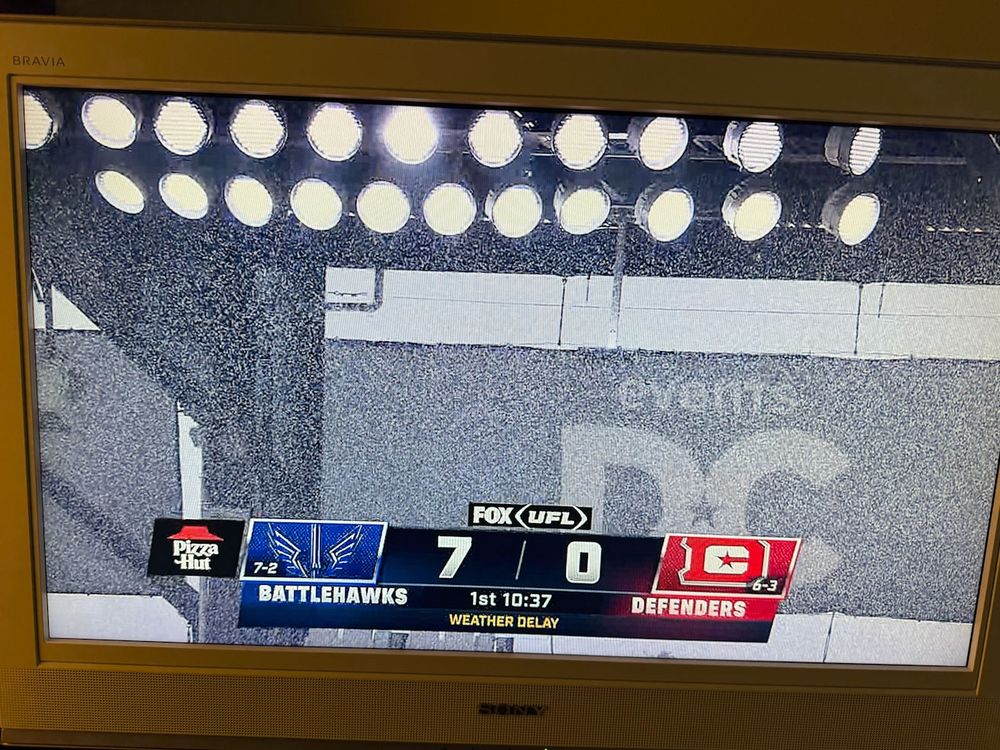 A television screen displaying a football game broadcast with a score of Battlehawks 7, Defenders 0. The game is under weather delay, with the time showing 10:37 in the first quarter. Bright stadium lights are visible above the scoreboard