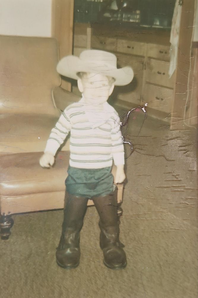 A scan of an old color photograph of a young boy. He is wearing adult cowboy boot that consume almost his entire legs, green pants and a long sleeve white t-shirt with horizontal green stripes. There is a very oversized tan cowboy hat on his head. The picture is faded and cracked by time but there is a big smile on the child's face that can just be made out.