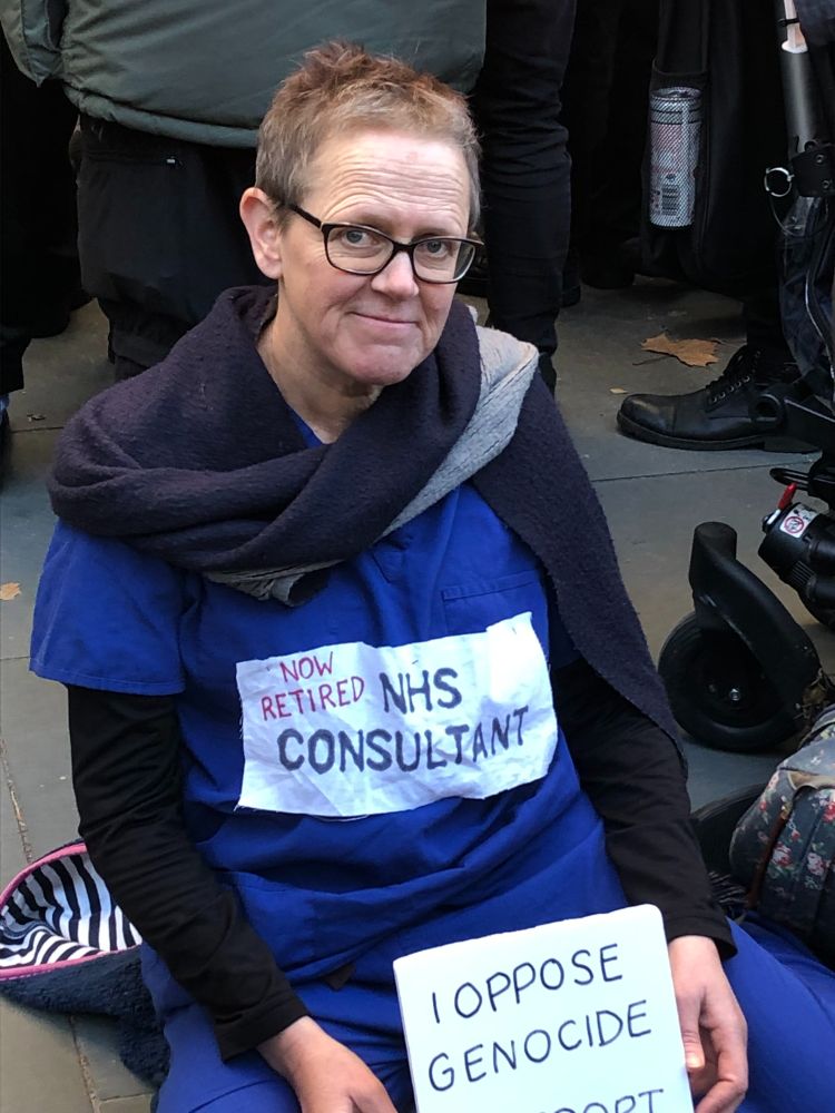 Older woman sitting on pavement, wearing scrubs with label "now retired NHS consultant" holding a sign "I oppose genocide. I support Palestine Action."