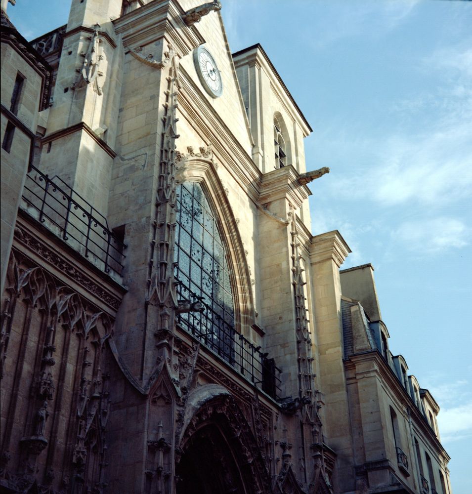 A color photograph of a Gothic church façade in Paris, featuring intricate stone carvings, a large stained glass window, and a clock tower bathed in warm evening light, taken with a Voigtländer Perkeo II and 80 mm f/3.5 Color-Skopar lens on Kodak Portra 160 film.