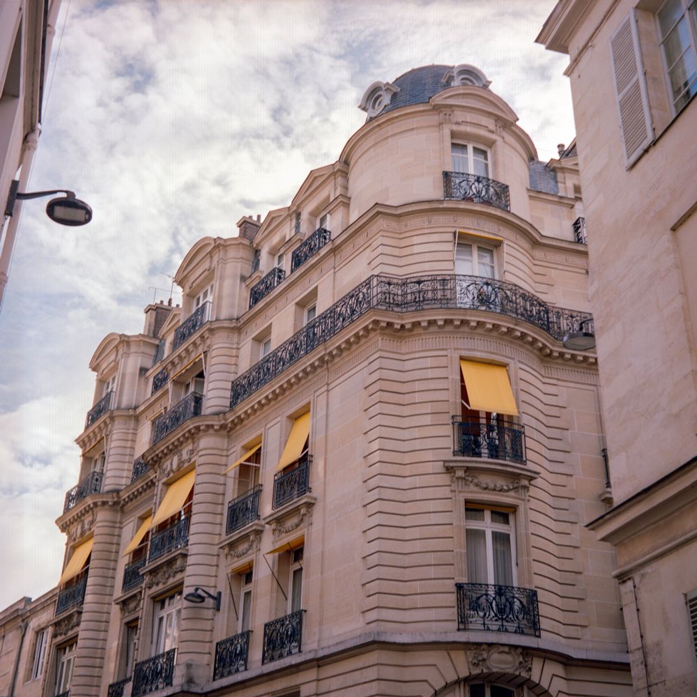 A soft-toned color photograph of a Haussmann-style Parisian building, captured with a Voigtländer Perkeo II and 80mm f/3.5 Color-Skopar lens on Kodak Portra 160. The pale cream stone facade is accented by wrought-iron balconies and bright yellow window awnings that stand out vividly against the pastel sky. 