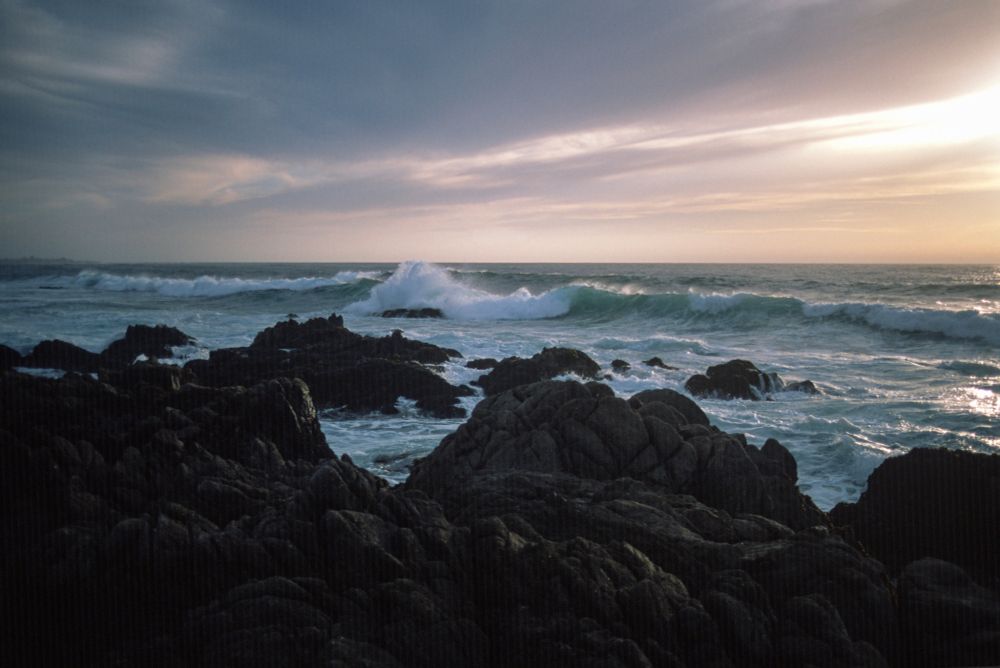 A color photograph of waves breaking against rugged coastal rocks beneath a soft, cloud-streaked sky. The ocean shimmers in cool blues and greens, with a single wave cresting dramatically near the center of the frame. The sun, partially obscured by thin clouds, casts a gentle glow across the horizon, blending golden and lavender hues. Captured on Fujicolor Provia 100 with a Pentax LX and 35mm f/2.8 lens, the image features fine detail, natural contrast, and the smooth tonal gradation typical of E6 slide film.