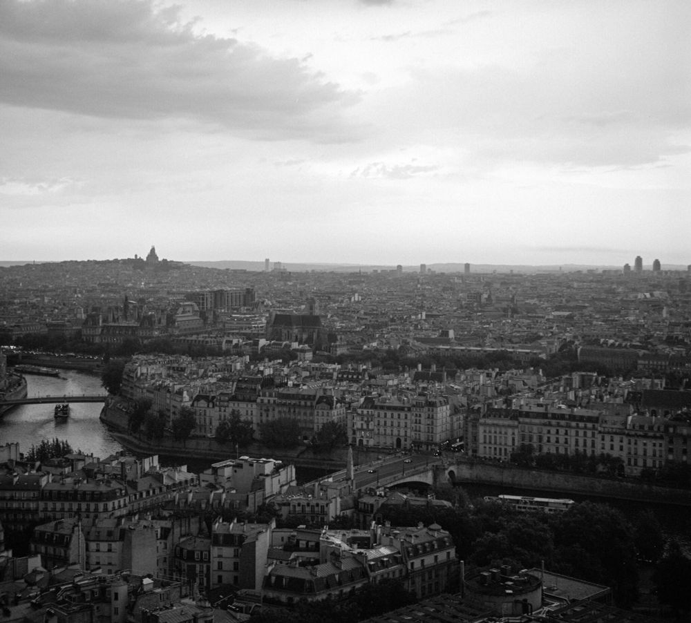 Black and white panoramic view of Paris at dusk, taken from a high vantage point. The Seine River curves through the city with boats visible, while the Sacré-Cœur Basilica stands silhouetted on the distant hill. The urban sprawl stretches toward the horizon under a moody, cloudy sky. Captured on a Voigtländer Perkeo II with an 80mm f/3.5 Color-Skopar lens using Ilford HP5+ film, developed in XTOL 1:1.