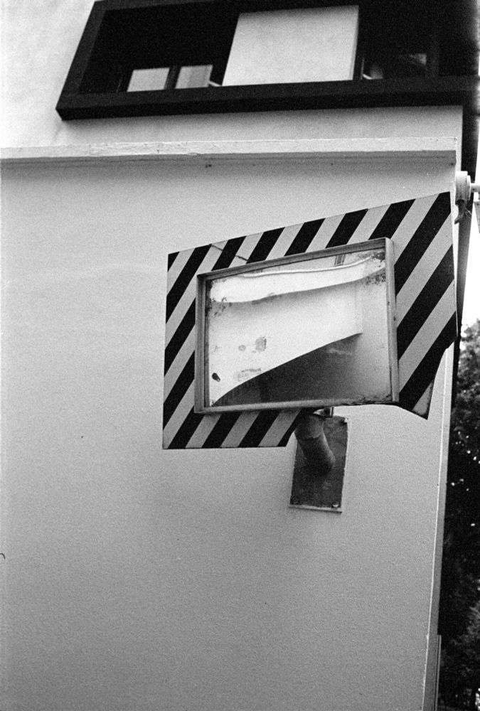 Black-and-white photo of a worn rectangular mirror with a striped caution border mounted on a wall, reflecting part of a building façade, captured with a Minolta CLE and Voigtländer Nokton Classic 35 mm f/1.4 lens on Kodak T-Max P3200 film, developed in XTOL 1:1.