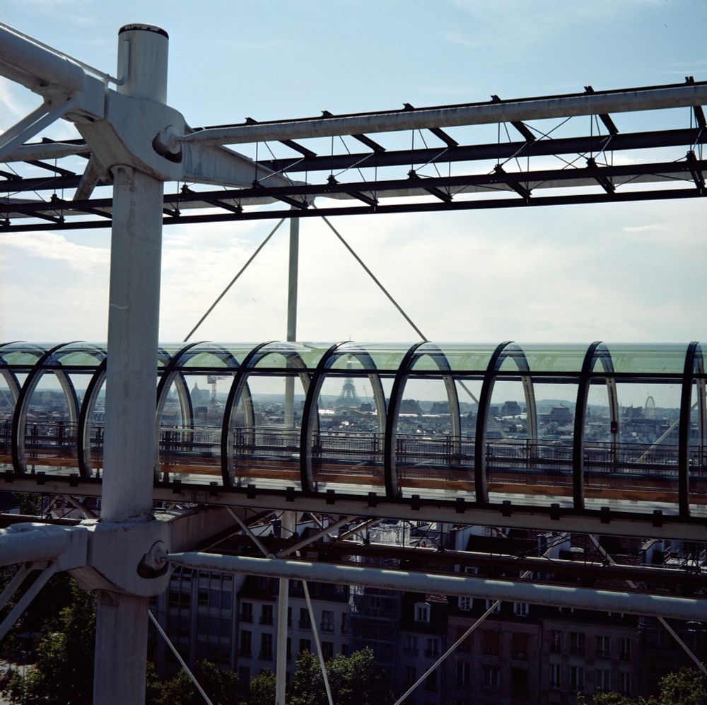 A color photograph of the Pompidou Centre’s iconic glass-enclosed escalator tube, elevated above Paris rooftops with the Eiffel Tower visible in the hazy distance, taken with a Voigtländer Perkeo II and 80 mm f/3.5 Color-Skopar lens on Kodak Portra 160 film.