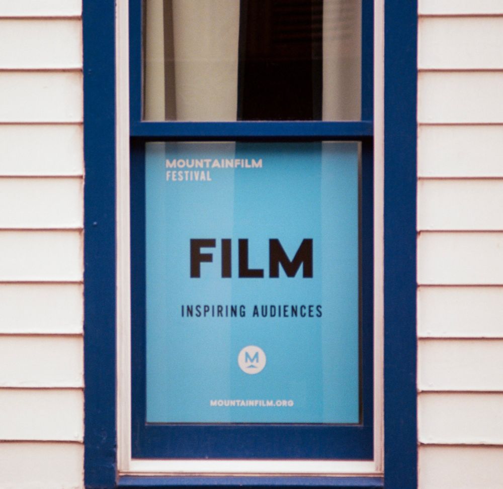 A color photograph of a window framed in blue on a white wooden building, displaying a blue poster for the Mountainfilm Festival. The poster reads “FILM — Inspiring Audiences” with the Mountainfilm logo and website below. Captured on Kodak Ektar 100 film using a Pentax MX with a 100mm f/2.8 lens.