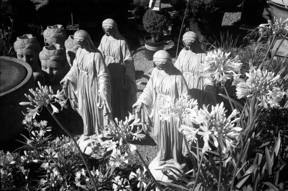 Black and white image of multiple robed religious statues arranged among blooming flowers and head-shaped planters in an outdoor garden center, taken with a LOMO Cosmic Symbol and T-43 40mm f/4 lens on Aviphot 200 film, developed in T-Max 1:4.