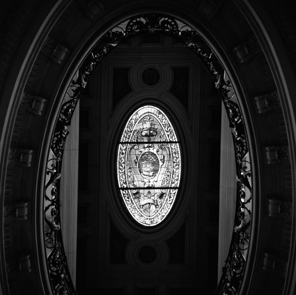  Black and white photograph looking upward through an oval opening framed by ornate wrought iron railings. At the center, a decorative oval stained-glass window glows with intricate patterns, floral motifs, and a crowned emblem. The surrounding architecture shows detailed moldings and classic design, emphasizing the symmetry and elegance of the interior. Captured on a Voigtländer Perkeo II with an 80mm f/3.5 Color-Skopar lens using Ilford HP5+ film, developed in T-Max 1:4.