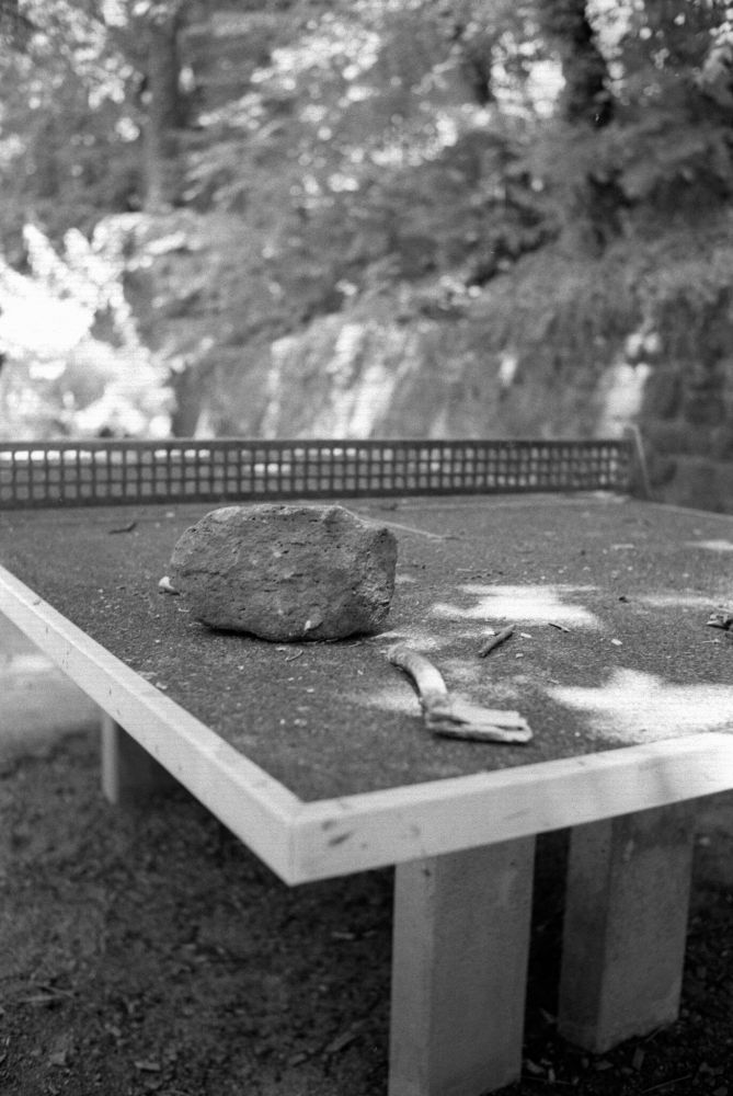 Black and white photo of an outdoor ping pong table covered in debris, including a large rock and several sticks, surrounded by dappled light from nearby trees; taken with a Minolta CLE and Voigtländer Nokton Classic 35mm f/1.4 lens using a red filter on Aviphot 200 film, developed in T-Max 1:4.