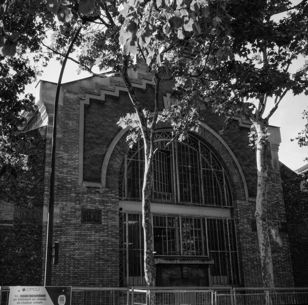 Black-and-white photo of a brick industrial building with a large arched window façade, partly obscured by tall trees, captured with a Voigtländer Perkeo II and 80 mm f/3.5 Color-Skopar lens on Kentmere Pan 200 film, developed in XTOL 1:1.