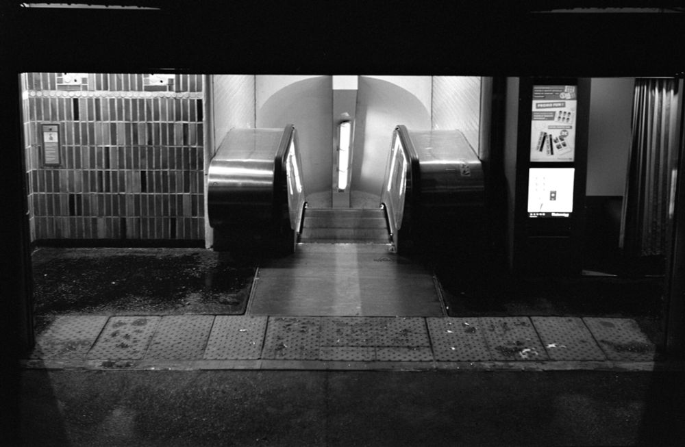 Black and white photo of a wet metro entrance with an illuminated escalator and tiled walls, shot on Voigtländer Perkeo II with 80mm f/3.5 Color-Skopar lens, Ilford HP5+ film developed in XTOL 1:1.