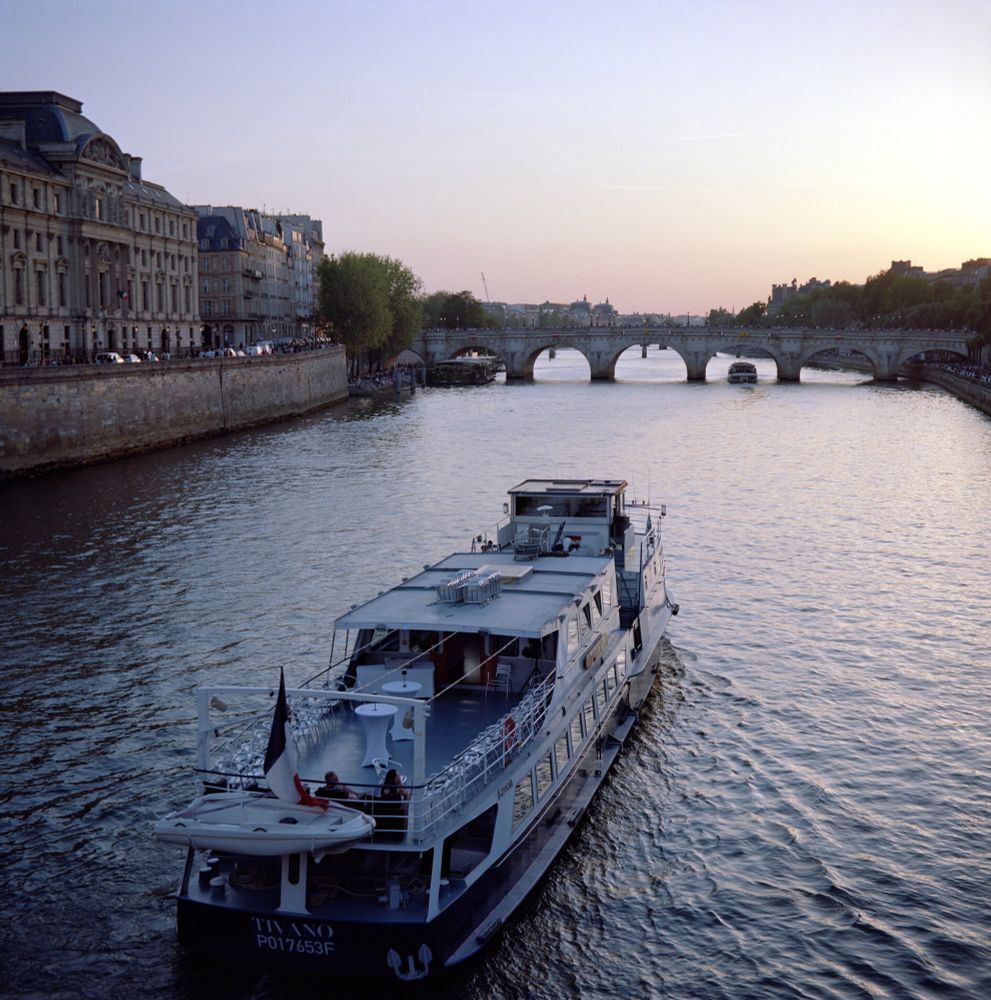 A serene evening scene on the Seine, with the riverboat Tivano gliding through the water beneath a fading pastel sky. The Pont Neuf stands in the distance, its arches reflecting the last light of sunset, while the riverbanks fill with life and activity. Captured with a Voigtländer Perkeo II on Kodak Portra 160 film, using the 80mm f/3.5 Color-Skopar lens.