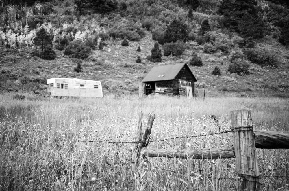 Black and white photograph of a rustic rural scene featuring an old wooden shack and a weathered trailer set against a hillside covered in grass and scattered trees. In the foreground, a rough wooden fence with barbed wire frames the image, its texture sharply rendered. The soft focus on the mid-ground adds depth, emphasizing the distance between the fence and the structures. Captured with an Olympus 35RC and 42mm f/2.8 E. Zuiko lens on Ilford XP2 Super 400 film, developed in the C-41 process.