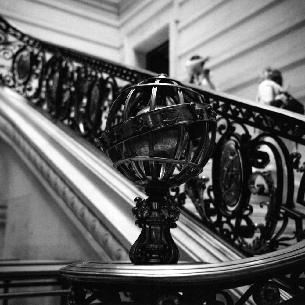 Black and white photograph of an ornate stairwell inside a grand building. The focus is on a decorative metal sphere mounted on the banister, resembling an armillary sphere. Behind it, the staircase railing features elaborate wrought iron designs. In the blurred background, two people can be seen ascending the stairs. Captured on a Voigtländer Perkeo II with an 80mm f/3.5 Color-Skopar lens using Ilford HP5+ film, developed in T-Max 1:4.