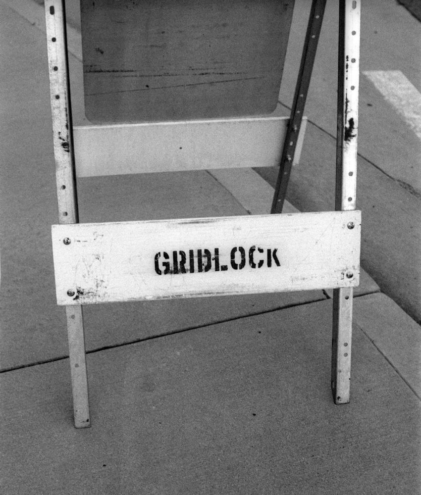 Black and white photo of a weathered street barricade with the word "GRIDLOCK" stenciled on a horizontal plank, taken with a Kodak Retina IIa and Schneider-Kreuznach Retina-Xenon 50mm f/2 lens on Aviphot 200 film, developed in T-Max 1:4.