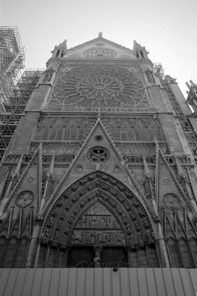 Black and white photo of the ornate Gothic façade of Notre-Dame Cathedral with its large rose window, scaffolding visible on both sides, shot on Minolta CLE with Voigtländer 21mm f/4 Color-Skopar lens using Kodak T-Max 100 film developed in T-Max (1:4).