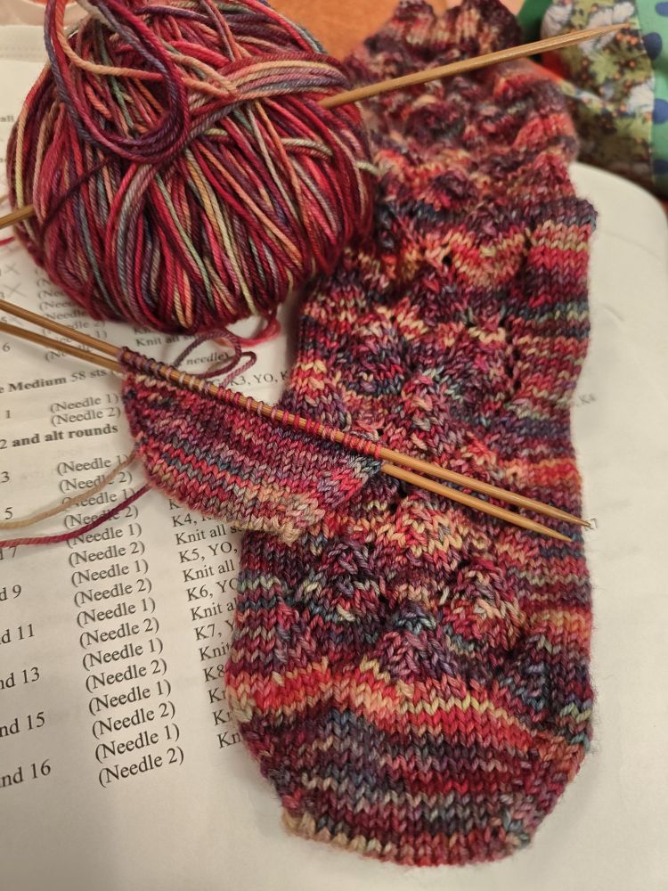 A multi coloured (reds, purples, and blues) hand knitted sock with a lace pattern on the front. Next to it is a ball of the yarn and the toe of the second sock. All on top of the paper pattern. 
