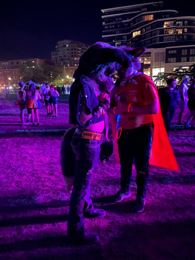 Two people in animal costume confer in an outdoor event space at night bathed in pinky purple stage light.